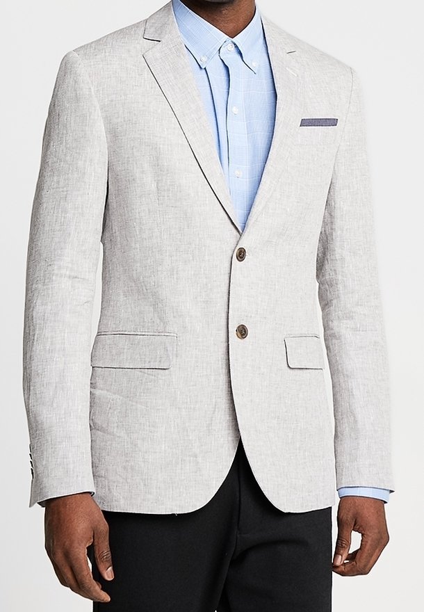 Homme portant un blazer gris clair à deux boutons sur une chemise bleu clair à col et un pantalon noir, debout devant un fond uni.