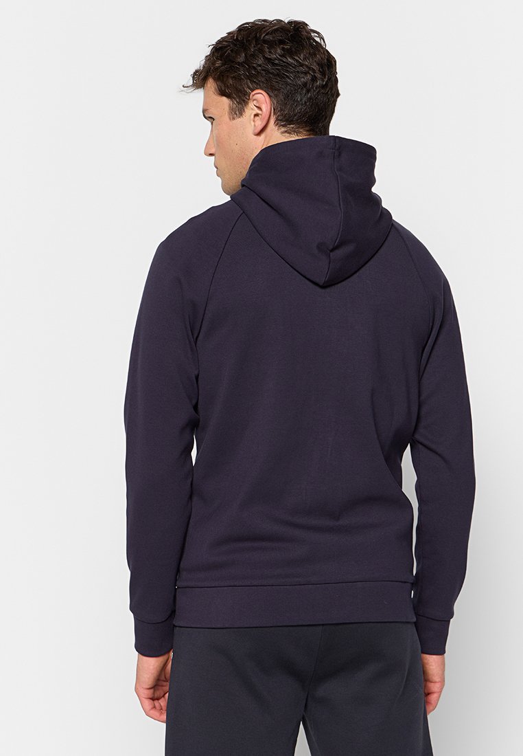 Uomo di spalle con indosso un felpa blu scuro con il cappuccio abbassato e pantaloni scuri abbinati, su uno sfondo bianco semplice.