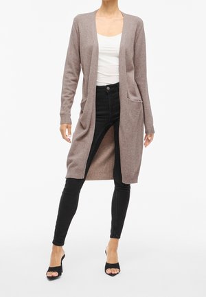 Femme portant un long cardigan beige ouvert devant avec des poches, un top blanc, un jean skinny noir et des sandales à talons ouvertes noires, debout les jambes écartées.