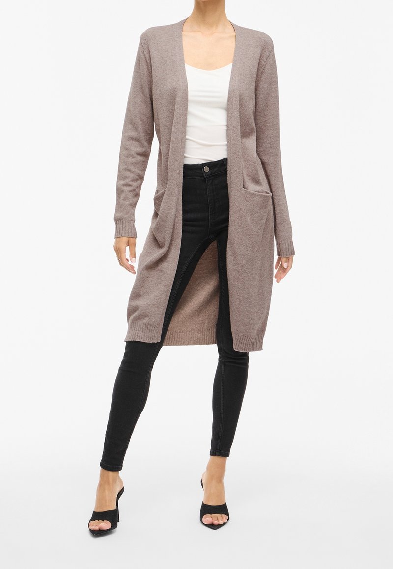Femme portant un long cardigan beige ouvert devant avec des poches, un top blanc, un jean skinny noir et des sandales à talons ouvertes noires, debout les jambes écartées.