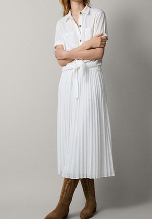 Robe midi plissée blanche à manches courtes, avec un devant à boutons et une ceinture à nouer, portée avec des bottes de style cowboy marron.