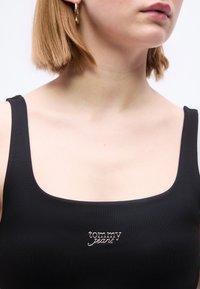 Femme aux cheveux blonds courts portant une boucle d'oreille en or en forme d'anneau et un débardeur noir avec le logo "tommy jeans" sur la poitrine.