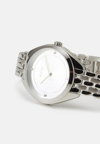 Montre en acier inoxydable argenté avec un boîtier rond, un cadran blanc, des index d'heure minimalistes et un design de bracelet lié. Présente une couronne sur le côté.