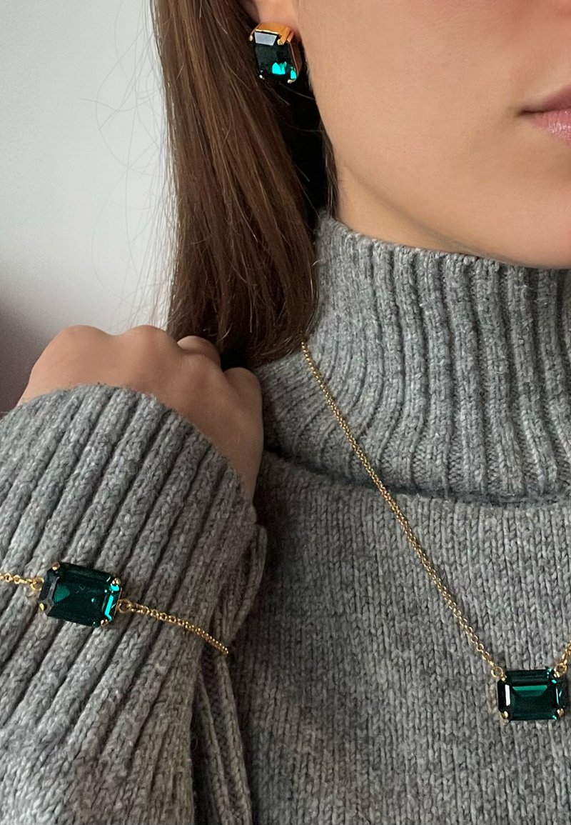 Smaragdgroene juwelenset, bestaande uit een geometrische oorbel en armband met gouden accenten, gedragen met een gestructureerde grijze coltrui.