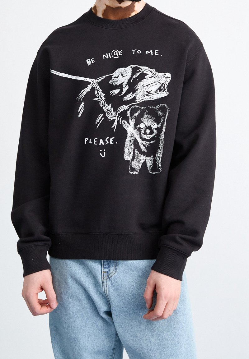Sudadera negra con un diseño gráfico de un oso y un perro, con texto en blanco que dice "SE BUENO CONMIGO. POR FAVOR." Tejido texturizado.