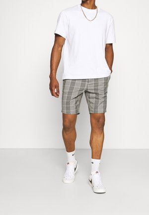 Camiseta blanca de algodón, pantalones cortos a cuadros grises con cintura elástica, llevados con zapatillas blancas y calcetines hasta el tobillo, de pie contra un fondo neutro.