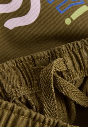 Olivově zelená látka s pasem na stažení; obsahuje barevný potisk textu v růžové a modré barvě. Texturovaný detail na pásku.