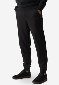 Pantalones de chándal negros de tela suave, con bolsillos laterales, puños elásticos y un logo sutil en el muslo. Combinados con zapatillas negras.