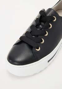 Svart lær sneakers med svarte snøringer, gull øyelets og hvit gummisåle. Har en rund tå og minimale sømdetaljer.