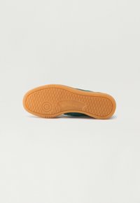 Gummisula av en grön Reebok-sneaker med texturerat grepp och synlig logotyp, fotograferad från botten mot en vit bakgrund.