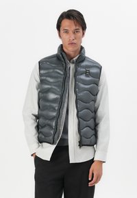 Gray quilted puffer vest with a high collar and zip closure. Features a small logo label near the chest and side pockets. Worn over a striped shirt.