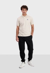 Polo beige à manches courtes, avec un petit logo coloré sur la poitrine, associé à un pantalon de jogging noir et des baskets blanches.