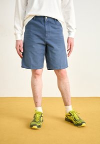 Blaue Shorts mit geradem Schnitt, mit einem vorderen Knopfverschluss und Taschen. Kombiniert mit grünen und gelben Sportschuhen.