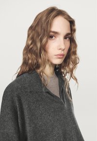 Maglione grigio lavorato a maglia con scollo a collo, texture morbida e vestibilità comoda, indossato sopra una maglietta grigio chiaro. Presenta capelli mossi che incorniciano il volto.