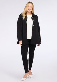 Manteau noir avec de grands boutons ronds, poches latérales et un col, associé à un haut blanc et un pantalon noir slim. Chaussures à talons blanches.