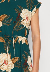 Vestido floral em tecido teal escuro com peónias brancas e pormenores castanhos. Tem mangas curtas e uma cintura ajustada com cordão.
