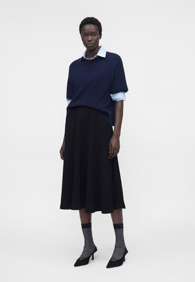 Γυναίκα που φοράει σκούρο μπλε πουλόβερ πάνω από ανοιχτό μπλε πουκάμισο, μαύρη midi φούστα, γκρι κάλτσες με μοτίβα και μαύρες μυτερές γόβες, στέκεται μπροστά σε λευκό φόντο.