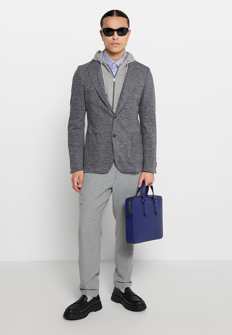 Mężczyzna w okularach przeciwsłonecznych, szary żakiet na szarym, zapinanym na zamek hoodie i niebieskiej koszuli, jasnoszare spodnie, czarne buty, trzymający niebieską teczkę.
