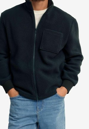 Mand iført en sort fleecejakke med lynlås og brystlomme samt blå jeans, hænderne i lommerne, foran en ensartet baggrund.