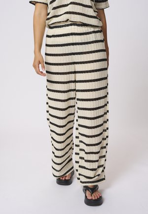 Pantalons larges à rayures noires et blanches en tissu tricoté texturé, avec une taille élastique et une coupe décontractée. Associés à des sandales noires.