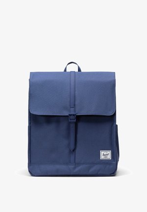 Zaino blu navy con chiusura a patta, design minimalista, cinturino regolabile con fibbia, manico superiore e una piccola etichetta con logo in basso.