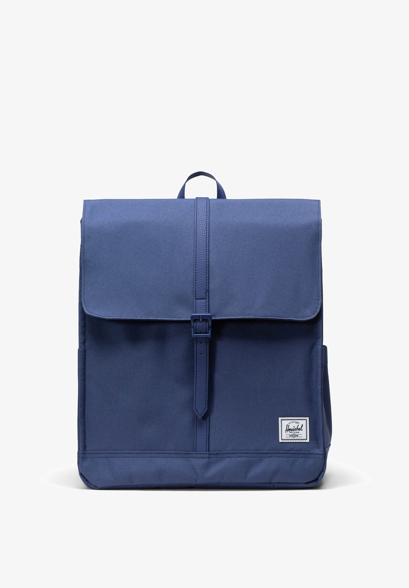 Zaino blu navy con chiusura a patta, design minimalista, cinturino regolabile con fibbia, manico superiore e una piccola etichetta con logo in basso.