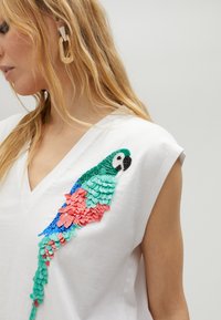 Top senza maniche in cotone bianco con un'applicazione di un pappagallo colorato in paillettes rosa, verde e blu sul lato sinistro, dotato di scollatura a V.