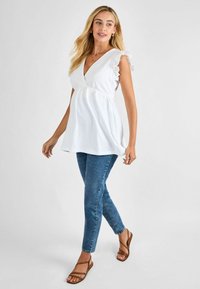 Weißes, strukturiertes V-Ausschnitt-Oberteil mit rüschigen Spitzenärmeln, kombiniert mit blauen Skinny-Jeans und braunen, geflochtenen Sandalen. Model läuft vor einem einfarbigen Hintergrund.