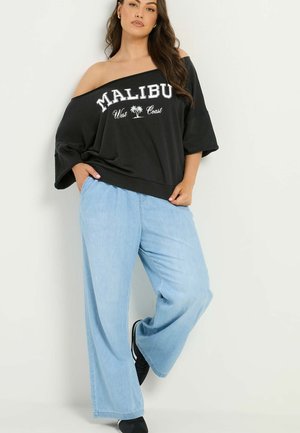 Kobieta w czarnym topie odkrywającym ramiona z tekstem "Malibu West Coast" i jasnoniebieskich szerokich dżinsach stoi na tle jednolitej ściany.