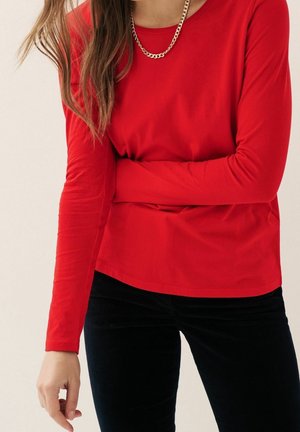 Femme portant un haut rouge à manches longues et un pantalon noir, accessoirisée d'un collier chaîne en or, debout devant un arrière-plan uni.
