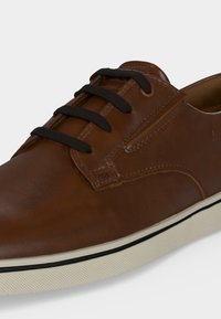 Chaussure décontractée en cuir marron avec lacets noirs et semelle en caoutchouc blanche avec bande noire, montrée en gros plan sous un angle.