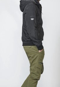 Homme portant une veste à capuche noire et un pantalon cargo vert olive, la main dans la poche, vu de profil sur un fond uni.