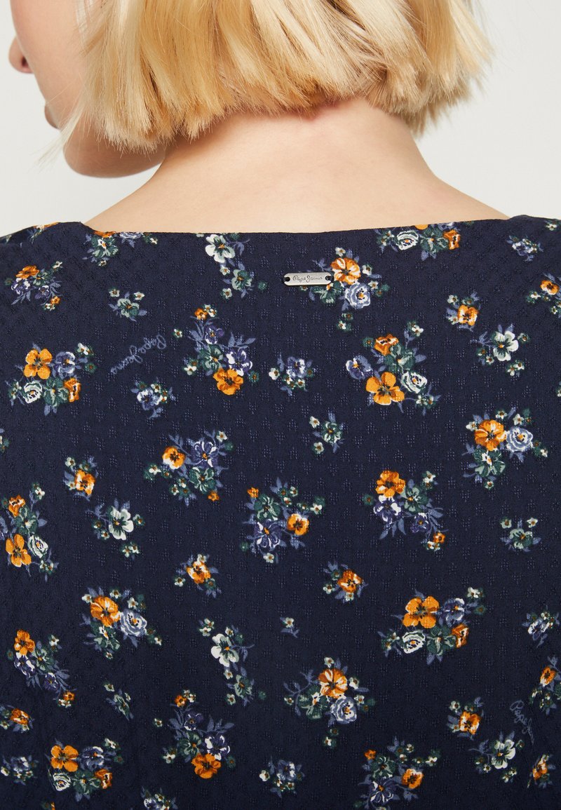 Persona bionda vestita con una maglia blu navy con piccoli motivi floreali arancioni, bianchi e viola, vista di spalle dalla parte superiore della schiena.