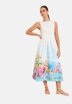 Woman wearing sleeveless white dress with colorful coastal and floral print skirt and white strappy sandals, standing with hand on hip.