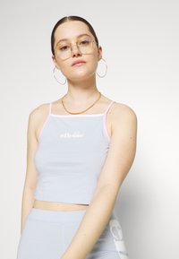 Ljusblå cropped tank top med rosa kant, med "ellesse"-logotypen i vitt. Slät tygtextur och tunna spaghettiband.