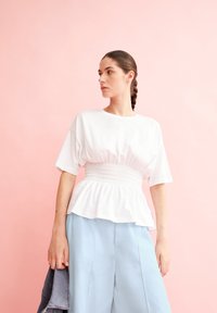 Witte katoenen top met een smocked taille en korte mouwen, gecombineerd met lichtblauwe wijde broek. Achtergrond is een zachte roze.