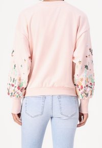 Felpa rosa chiaro con maniche a palloncino, caratterizzata da un vivace ricamo floreale. Completata con un orlo a coste, abbinata a jeans azzurri.