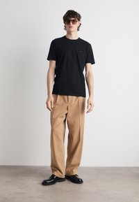 Čierne bavlnené tričko s malým logom, spárované s voľnými béžovými nohavicami a čiernymi robustnými topánkami. Model nosí tmavé slnečné okuliare.