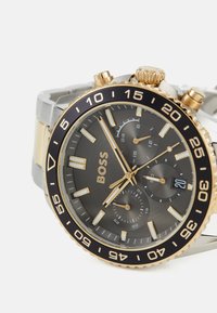 Montre analogique pour homme avec un cadran gris, des accents dorés, un bracelet en acier inoxydable argenté, une lunette noire et des sous-cadrans de chronographe.