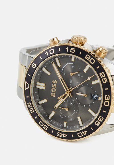 Montre analogique pour homme avec un cadran gris, des accents dorés, un bracelet en acier inoxydable argenté, une lunette noire et des sous-cadrans de chronographe.