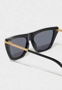 Óculos de sol estilo gato pretos com lentes escuras e braçadeiras de metal dourado gravadas com "ALLSAINTS" sobre um fundo branco.
