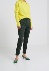 Person klädd i en ljusgul tröja, rutiga gröna och svarta byxor samt låga skor med gröna spetsiga tår som står på ett vitt golv.