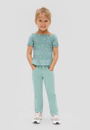 Mintgrünes geschmücktes Oberteil mit kurzen Rüschärmeln, kombiniert mit mintgrünen Hosen. Weiße und graue Sneakers. Blumenmuster auf dem Oberteil.