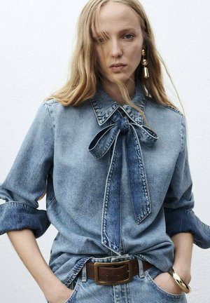Donna con capelli lunghi biondi che indossa una camicia di denim arrotolata con dettaglio a cravatta, jeans di denim, cintura marrone, orecchino d'oro e bracciale.