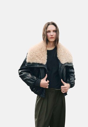 Chaqueta bomber de cuero negro con un gran cuello de borrego color beige, combinada con un suéter oscuro y pantalones de pierna ancha verdes.