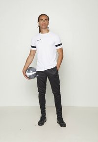 Weißes Nike-T-Shirt mit kurzen Ärmeln und schwarzen Akzenten, kombiniert mit schwarzen Jogginghosen und schwarzen Schuhen, hält einen silbernen Ball und steht vor einem schlichten Hintergrund.