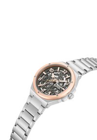 Reloj de acero inoxidable plateado con diseño esquelético, bisel en oro rosa y esfera gris. Presenta números grandes y una pulsera de eslabones de metal.