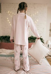 Valkoinen pitkähihaine paita yhdistettynä vaaleanpunaisiin pyjamahousuihin, joissa on karkkikeppikuviot. Vuoteessa on raidallisia ja yksivärisiä tyynyjä.