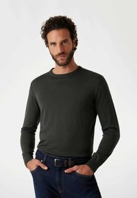 Dunkelgrüner Pullover mit langen Ärmeln und rundem Ausschnitt, getragen zu dunkelblauen Jeans und einem geflochtenen marineblauen Gürtel. Der Stoff erscheint glatt und figurbetont.