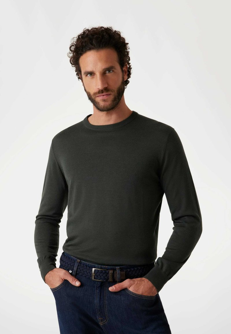 Dunkelgrüner Pullover mit langen Ärmeln und rundem Ausschnitt, getragen zu dunkelblauen Jeans und einem geflochtenen marineblauen Gürtel. Der Stoff erscheint glatt und figurbetont.